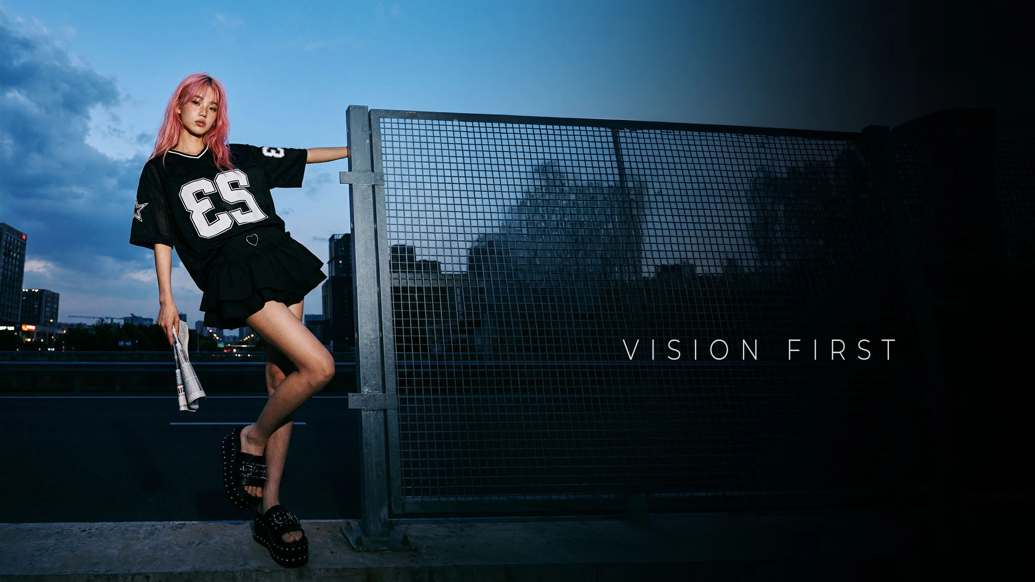 Vision First — editorial fashion rooftop, recinzione a maglie e skyline al crepuscolo